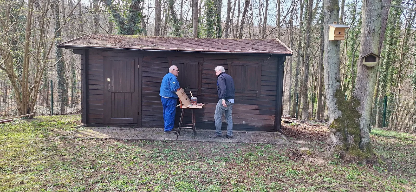 Zwei Mitglieder mit Werkzeug und Farbe vor dem hölzernen Geräteschuppen.